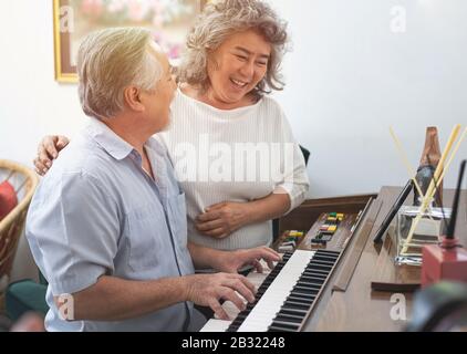 Anziano anziano uomo suona piano in casa di cura ascoltato da donna anziana, ritrattamento anziani nonna asiatica e nonno suonare piano in casa Foto Stock