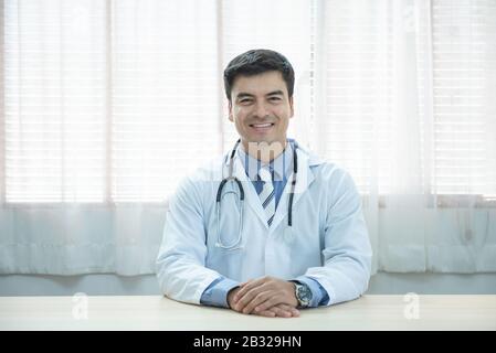 Giovane dottore caucasico seduto alla scrivania al suo posto di lavoro e sorridente guardando la macchina fotografica. Servizio medico perfetto in clinica. Felice futuro di Foto Stock