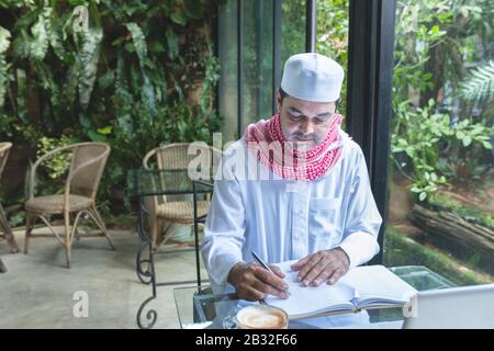 Giovane e bel uomo d'affari arabo musulmani in caffetteria indossando hijab vestito scrivere su libro file con portatile sul tavolo. Foto Stock