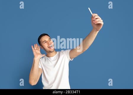 Prendere selfie. Ritratto del giovane caucasico isolato su sfondo blu studio. Bel modello maschile in stile casual, colori pastello. Concetto di emozioni umane, espressione facciale, vendite, pubblicità. Foto Stock