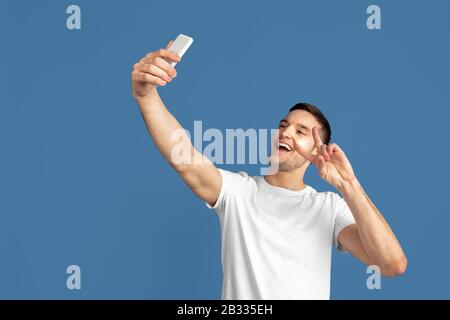 Prendere selfie. Ritratto del giovane caucasico isolato su sfondo blu studio. Bel modello maschile in stile casual, colori pastello. Concetto di emozioni umane, espressione facciale, vendite, pubblicità. Foto Stock