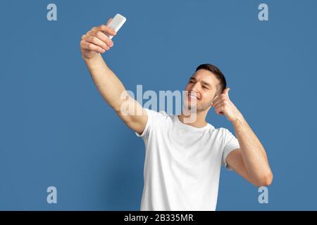 Prendere selfie. Ritratto del giovane caucasico isolato su sfondo blu studio. Bel modello maschile in stile casual, colori pastello. Concetto di emozioni umane, espressione facciale, vendite, pubblicità. Foto Stock