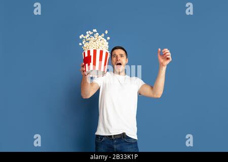 Cinema emozionante guardare, pop corn volare. Ritratto del giovane caucasico su sfondo blu studio. Modello maschile in stile informale, colori pastello. Concetto di emozioni umane, espressione facciale, vendite, pubblicità. Foto Stock