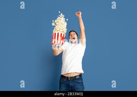 Cinema emozionante guardare, pop corn volare. Ritratto del giovane caucasico su sfondo blu studio. Modello maschile in stile informale, colori pastello. Concetto di emozioni umane, espressione facciale, vendite, pubblicità. Foto Stock