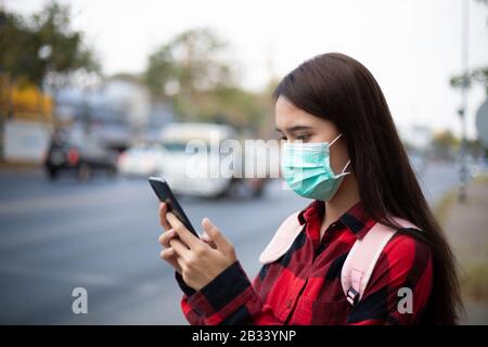 Asian giovane donna che usa lo smartphone in città indossando maschera viso a causa di inquinamento atmosferico, particolato e per la protezione del virus influenzale, influenza, coro Foto Stock