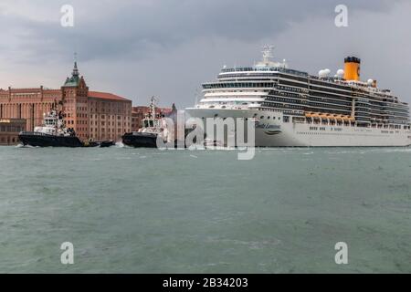 Due Tug barche tirare la massiccia nave da crociera, Costa Luminosa, che domina lo skyline di Venezia, italia, come si lascia in serata. Foto Stock