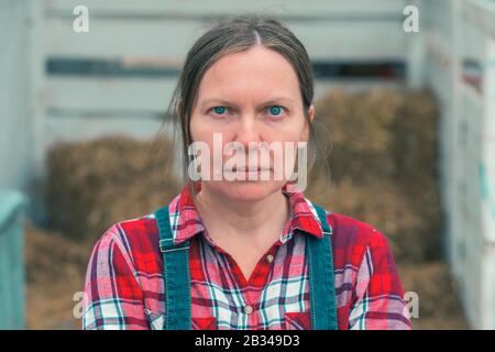 Grave interessato femmina agricoltore che posa in azienda. Donna lavoratore sicuro che indossa la camicia in plaid e jeans tute guardando la fotocamera. Foto Stock