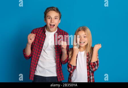 Ragazzo e ragazza eccitati celebrano il successo, alzando i pugni su sfondo blu Foto Stock