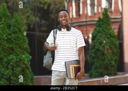 Felice ragazzo africano studente con zaino e libri di lavoro su sfondo universitario Foto Stock