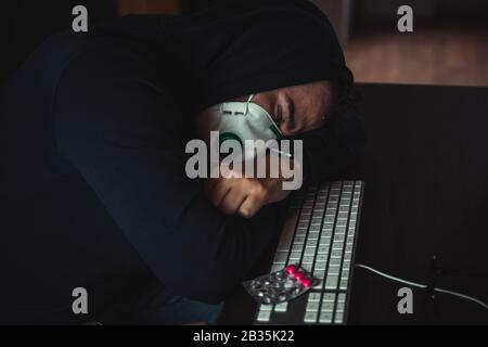 Uomo infettato da virus corona che dorme sulla tastiera al lavoro, maschera respiratore sul viso Foto Stock