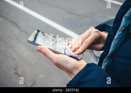 Mani femminili che strofinano lo schermo del telefono cellulare con tessuto antibatterico. Donna che pulisce il suo smartphone in piedi all'aperto, primo piano. Foto Stock