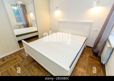 camera da letto in colori bianchi nella casa. Grande letto matrimoniale bianco, grande specchio sul guardaroba scorrevole, pavimento in parquet beige. Foto Stock