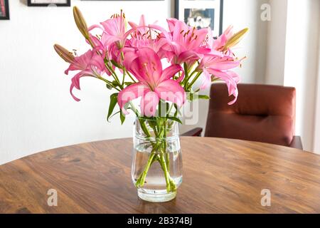 Bella gigli rosa fresco con una brocca di vetro come vaso su un bel tavolo di acacia in legno con una poltrona in pelle di cognac contro una parete bianca con... Foto Stock