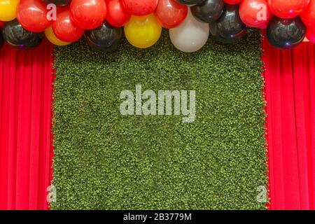 Bella parete verde erba pannello con palloncini colorati sopra e tenda rossa sui lati. Spazio di copia. Area di testo. Foto Stock