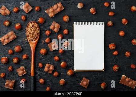 Cioccolato con nocciole, un taccuino con pagine bianche e un cucchiaio di legno con cacao su sfondo scuro, circondato da noci nel guscio e sbucciato. Foto Stock