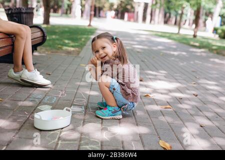 Carina bambina seduta e disegno con gesso su asfalto nel parco. Foto Stock
