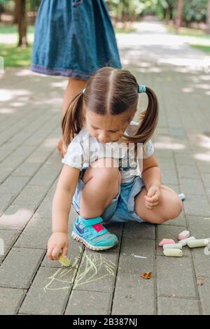 Carina bambina seduta e disegno con gesso su asfalto nel parco. Foto Stock