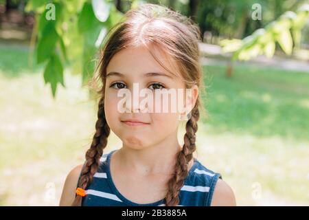 Ritratto di una bambina carina sconvolta con i pigtail in piedi e guardando la fotocamera in estate Foto Stock