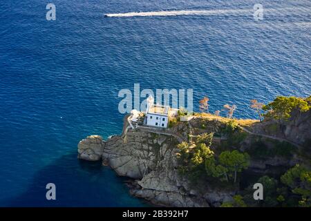 Il faro bianco si trova in cima alla collina del Mar Ligure, con la barca a vela bianca sullo sfondo. Arial vista sulla costa rocciosa verde in Foto Stock