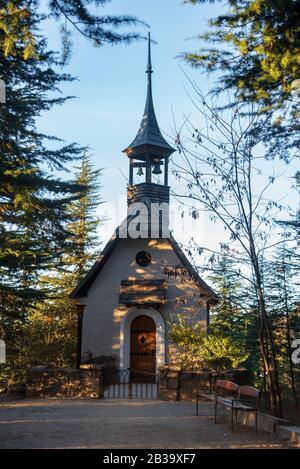 Piccola chiesa germanica in legno con due campane alla torre di la Cumbrecita, con una strada sterrata e una panca di fronte ad essa, circondata da tr sempreverde Foto Stock