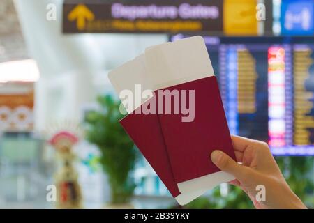 Due passaporti e carte d'imbarco in mano di una donna di fronte al cartello informativo per la partenza e l'arrivo all'interno di un aeroporto. Foto Stock