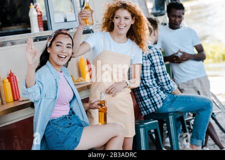 fuoco selettivo ragazza asiatica felice che ondola mano vicino a donna redhead e camion del cibo Foto Stock