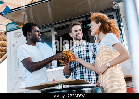 vista a basso angolo di uomini multiculturali felici bottiglie di birra con attraente redhead donna vicino al camion alimentare Foto Stock