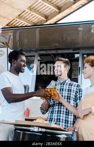 focus selettivo di uomini multiculturali che graffano bottiglie di birra con felice redhead donna vicino al camion alimentare Foto Stock