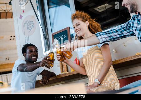 bei uomini multiculturali che graffano le bottiglie di birra con attraente donna rossa vicino al camion del cibo Foto Stock