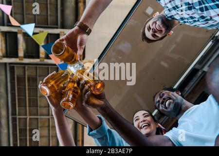 vista dal basso di amici multiculturali felici tostare bottiglie di birra Foto Stock
