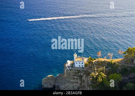 Il faro bianco si trova in cima alla collina del Mar Ligure, con la barca a vela bianca sullo sfondo. Arial vista sulla costa rocciosa verde in Foto Stock