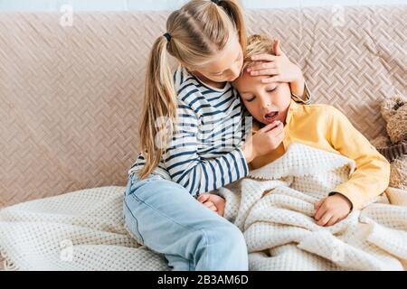 sorella attenta che dà la pillola al fratello malato e che tocca la fronte Foto Stock