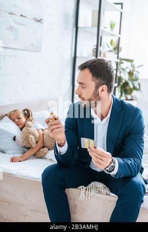 padre che tiene pillole e spray nasale mentre si siede a letto vicino a figlia ammalata Foto Stock