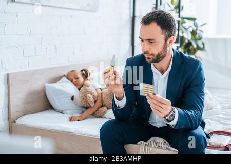 il padre serio che tiene le pillole e lo spruzzo nasale mentre si siede sul letto vicino alla figlia ammalata Foto Stock