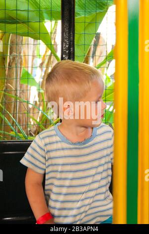 Un bambino di tre anni fa si muove in un trenino verde-giallo per bambini in un parco divertimenti in Riviera, Sochi, di fronte allo spettatore. Riposo, tropici, riposo, attivazione. Foto verticale Foto Stock