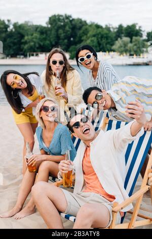 allegro giovane seduto in chaise lounge e prendere selfie con sorridenti amici multiculturali sulla spiaggia Foto Stock