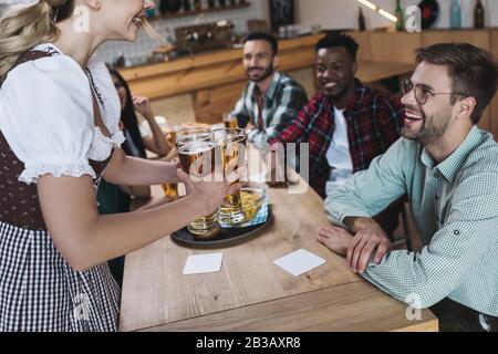 vista corta della cameriera che tiene bicchieri di birra leggera vicino ad amici multiculturali Foto Stock