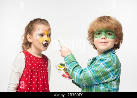 allegro ragazzo con maschera gecko dipinta che tiene tavolozza e pennello vicino amico con muso tigre dipinto sul viso isolato su bianco Foto Stock