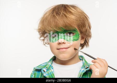 cute boy con maschera gecko dipinta su guancia toccante viso con pennello isolato su bianco Foto Stock