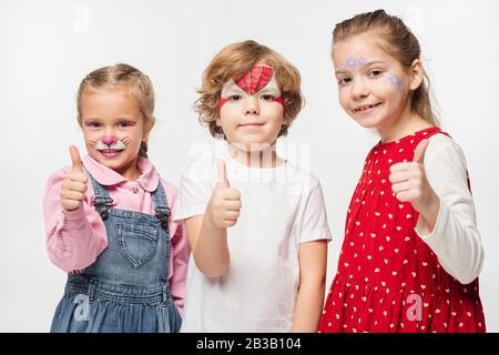 amici allegri con dipinti colorati che mostrano i pollici mentre si guarda la fotocamera isolata su bianco Foto Stock