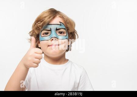 ragazzo allegro con maschera batman dipinta sul viso che mostra il pollice in alto guardando la fotocamera isolata su bianco Foto Stock