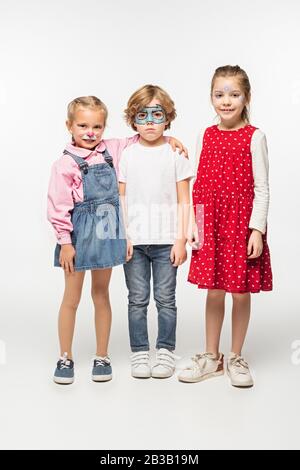 vista a tutta lunghezza di bambini allegri e ragazzo offeso con dipinti colorati che guardano la macchina fotografica su sfondo bianco Foto Stock