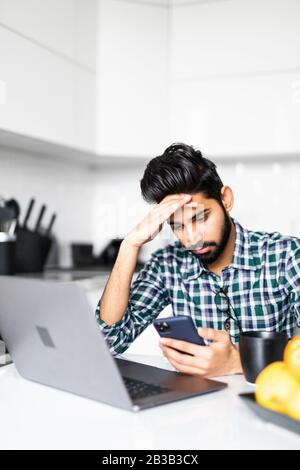 Stanco indiano uomo che tiene il telefono che lavora sul computer portatile al tavolo a casa, bere caffè in cucina Foto Stock