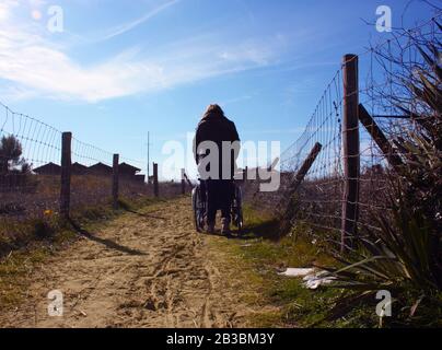 persona solitaria da dietro che spinge una sedia a rotelle da sola lungo un sentiero sabbioso che conduce al mare nelle dune della toscana Foto Stock