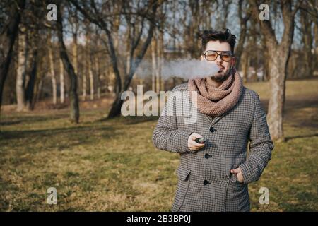 Sicuro giovane maschio con bearded in occhiali da sole vape sigaretta elettronica. Un giovane e bell'ippopotere che vapica e-sigaretta all'aperto nel parco Foto Stock