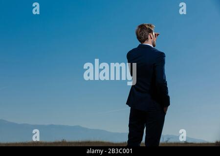 Vista posteriore di un uomo d'affari desideroso che guarda lontano mentre indossa un vestito blu, camminando sulle montagne esterne e sullo sfondo del campo Foto Stock