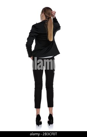 vista posteriore di una donna d'affari preoccupata indossando vestito nero e in piedi con la mano in tasca mentre graffia la testa contro sfondo bianco studio Foto Stock