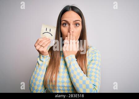 Giovane donna bionda che tiene la nota appiccicosa della carta con emoticon triste del viso sopra la bocca di copertura isolata dello sfondo con la mano scioccata con la vergogna per errore, exp Foto Stock