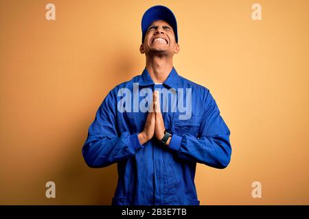 Giovane meccanico afroamericano che indossa uniforme blu e cappellino su sfondo giallo accattando e pregando con le mani insieme con l'espressione di speranza o Foto Stock