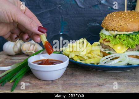 Primo piano hamburger su un piatto blu, accanto a cipolle tritate, patatine fritte e ketchup. La mano prende 1 patatine fritte impregnate in salsa Foto Stock